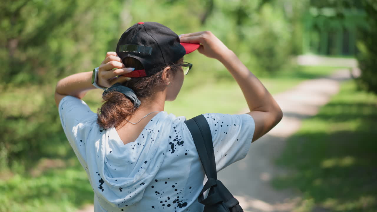 Persona caminando por el sendero de un parque con sudadera y mochila al hombro, ajustándose la gorra bajo la luz moteada del sol, vegetación veraniega enmarcando el camino, vista trasera que captura un tranquilo desplazamiento y una reflexión serena