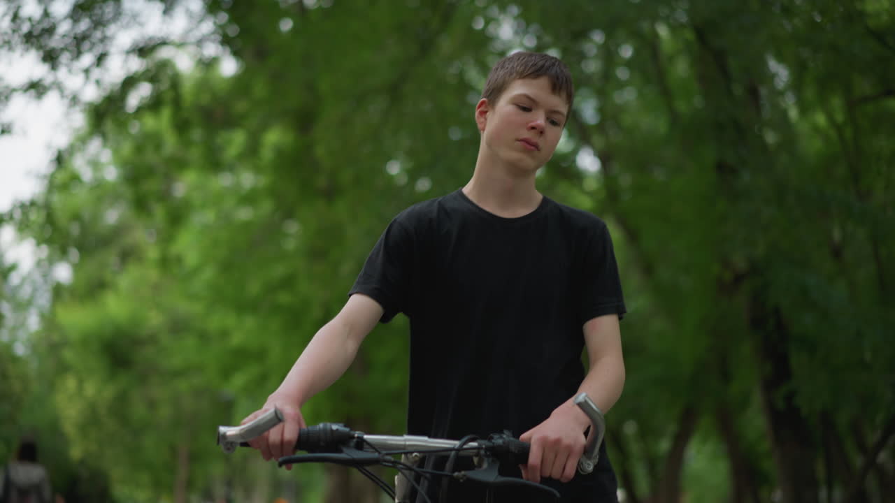 검은 옷을 입은 어린 소년은 평화로운 나무로 둘러싸인 길에서 자전거와 함께 걸어가고, 배경은 부드럽게 흐릿하며, 초록색 가운데 멀리서 두 사람을 보여줍니다.