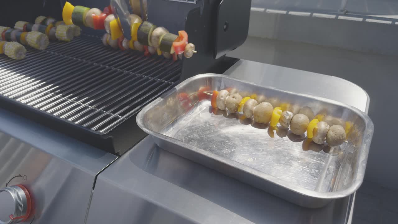Vegetable skewers with corn, pepper, zucchini, potatoes and mushrooms are placed on a stainless steel bbq using stainless steel tongs