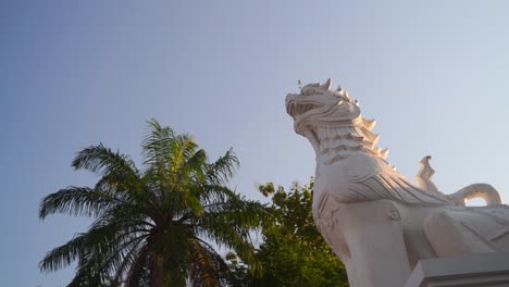 Mirando-Hacia-La-Estatua-Del-León-Dragón-Blanco-Contra-El-Cielo-Azul-Con-Palmera