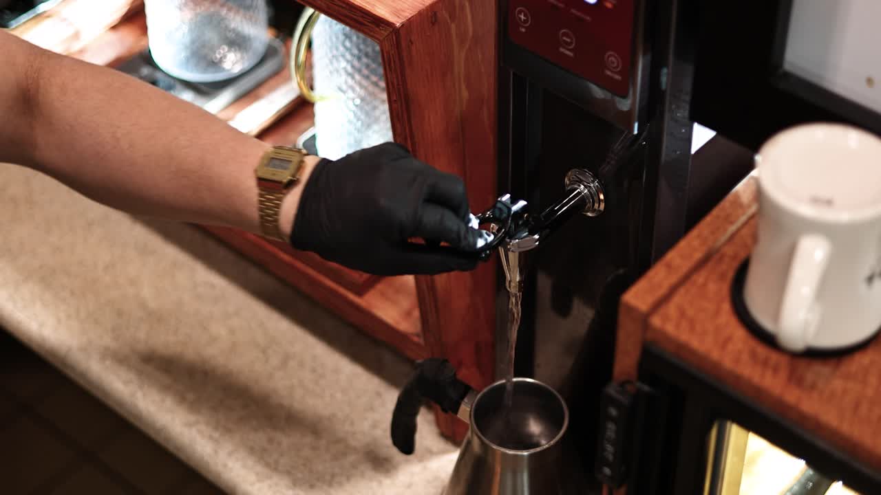 Neddykkende optagelse af en barista, der placerer en kedel under en varmtvandstud og fylder den – rustfri kande, jævn stråle, blød bokeh og detaljeret, ren arbejdsgang på caféen