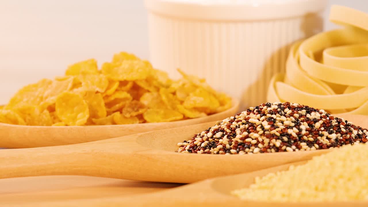 A close-up view of various grains, pasta, and cornflakes arranged on a wooden surface with bright lighting