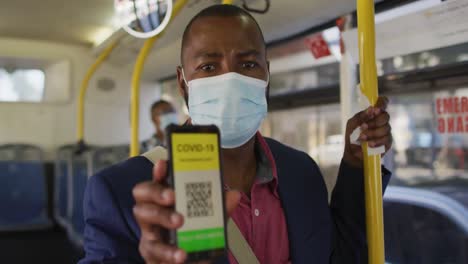 african american man wearing face mask, using smartphone with covid passport in bus