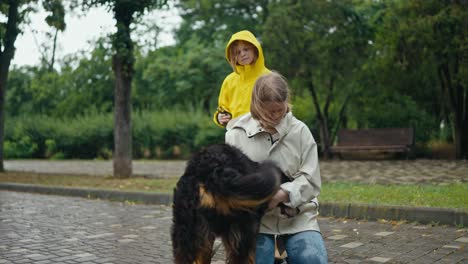 Glückliche-Blonde-Frau,-Die-Spaß-Hat-Und-Mit-Ihrem-Großen-Schwarzen-Haushund-Spielt,-Während-Sie-Nach-Dem-Regen-Mit-Ihrer-Kleinen-Tochter-Durch-Die-Gasse-Im-Park-Geht