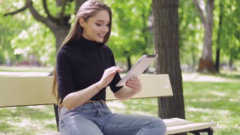 Smiling-beautiful-woman-sitting-on-bench-in-green-park-and-using-tablet