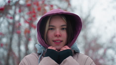 joven sonriendo ligeramente a la cámara, con chaqueta de invierno con capucha, al aire libre, fondo borroso con árboles y nieve, retrato de invierno casual, clima frío, entorno de parque urbano