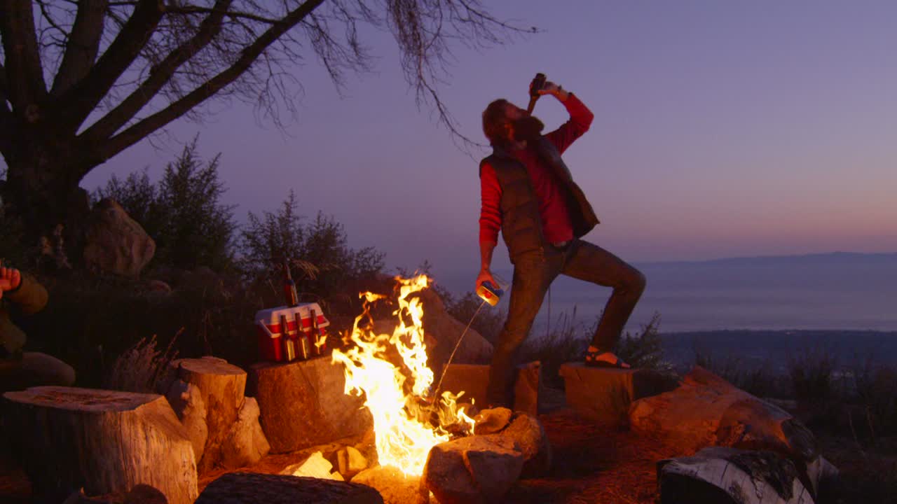 한 남자가 캠프장에서 맥주를 마시면서 모닥불에 라이터 액체를 뿌린다