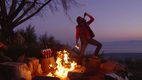 a man sprays lighter fluid on a campfire while drinking beer at a campsite