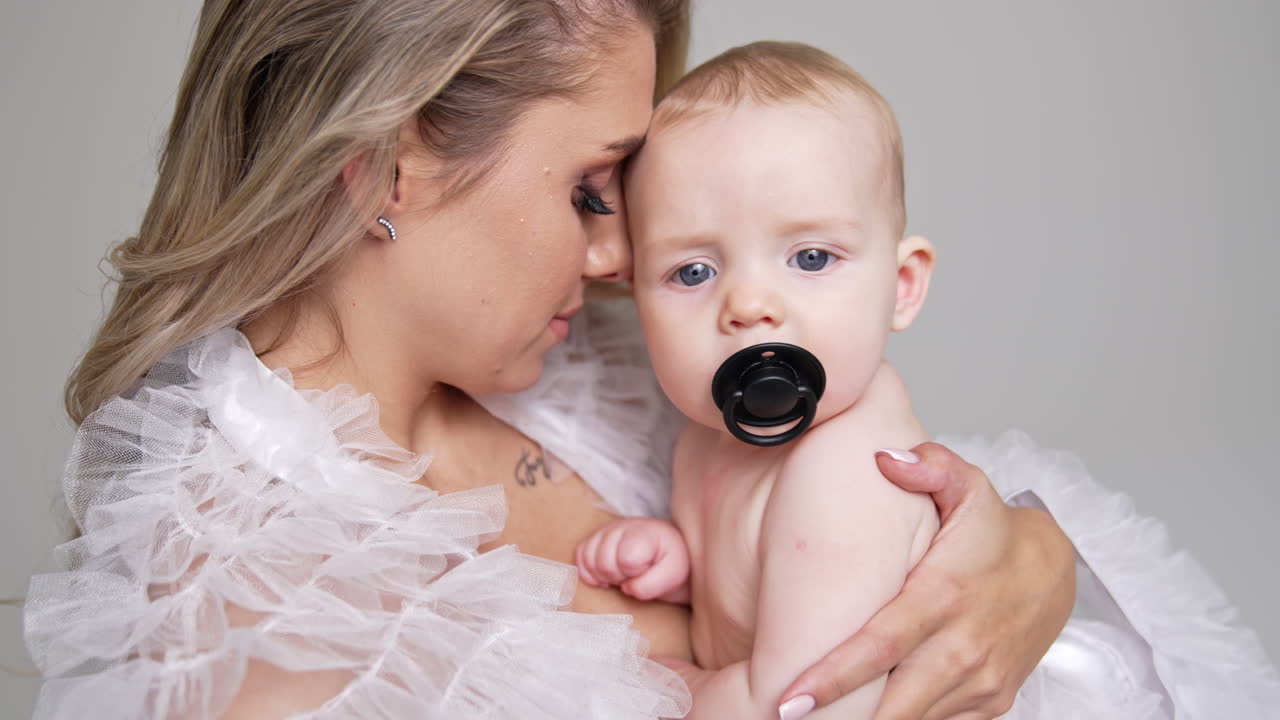 Woman holds her bare baby boy sucking a pacifier. Mother presses her beloved son to the heart. White backdrop. Close up.