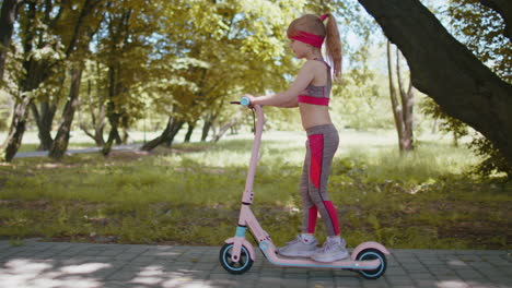 Young-athletic-fit-blonde-girl-rides-traveling-on-electric-scooter-on-road-in-park-on-sunny-day