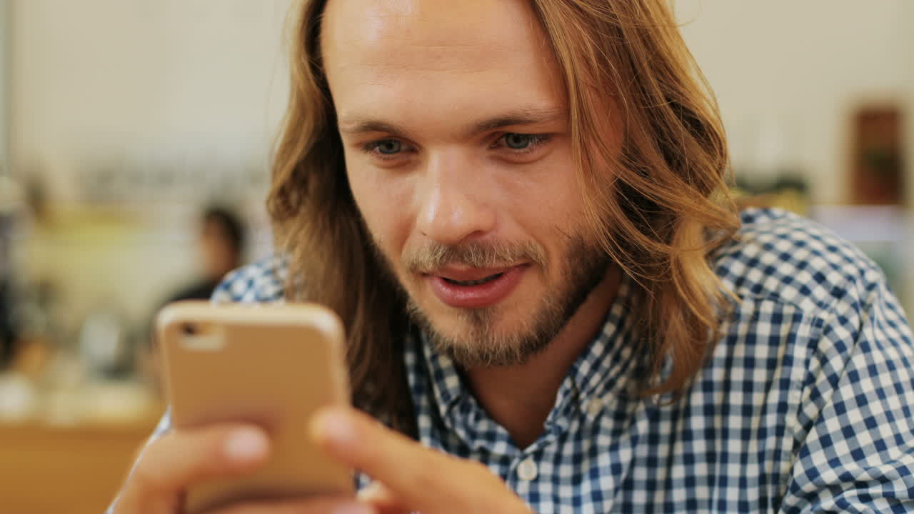 vista de cerca de un hombre rubio caucásico con el pelo largo enviando mensajes de texto por teléfono sentado en una mesa en un café