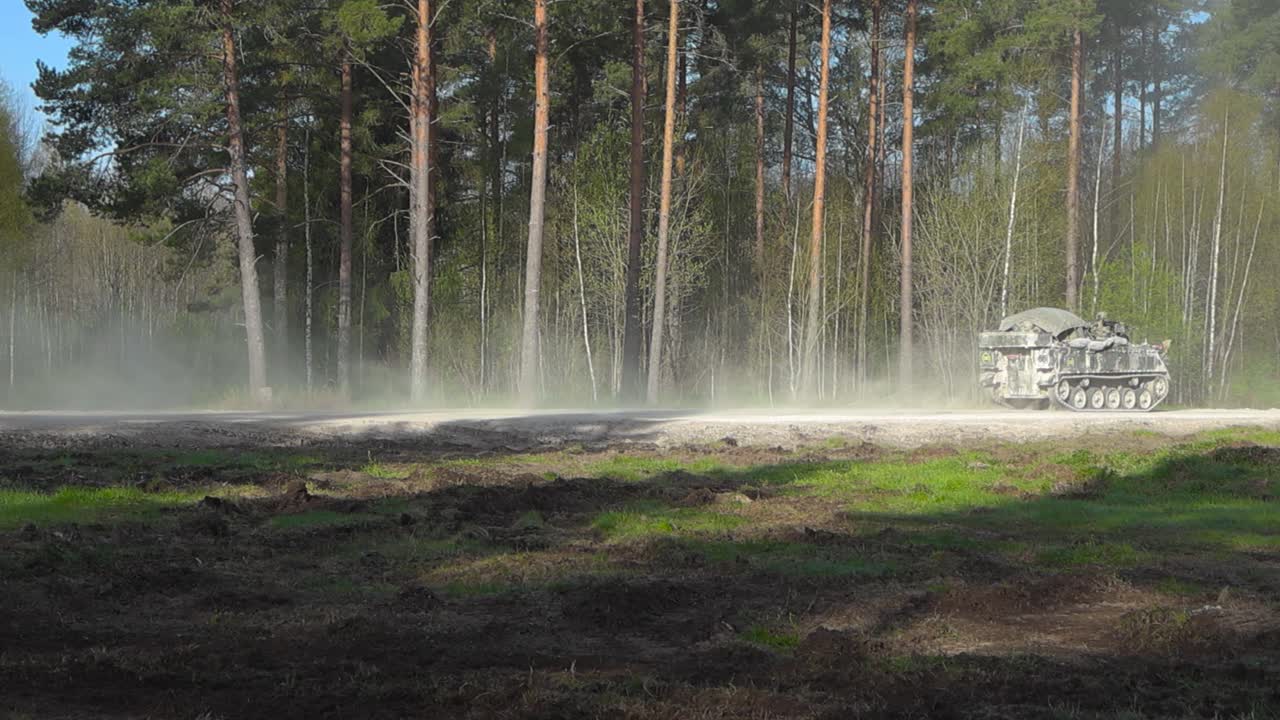 British military Bulldog army armoured personnel carrier driving on a gravel dry road in a sunny forest with a massive dust cloud behind it due to high speeds. Armed soldiers are on top of the tank.