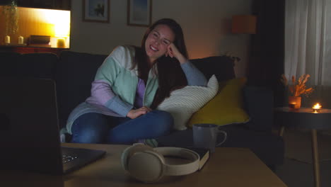 mujer sonriente sentada en el sofá en casa por la noche transmitiendo o viendo una película o un programa en la computadora portátil 6