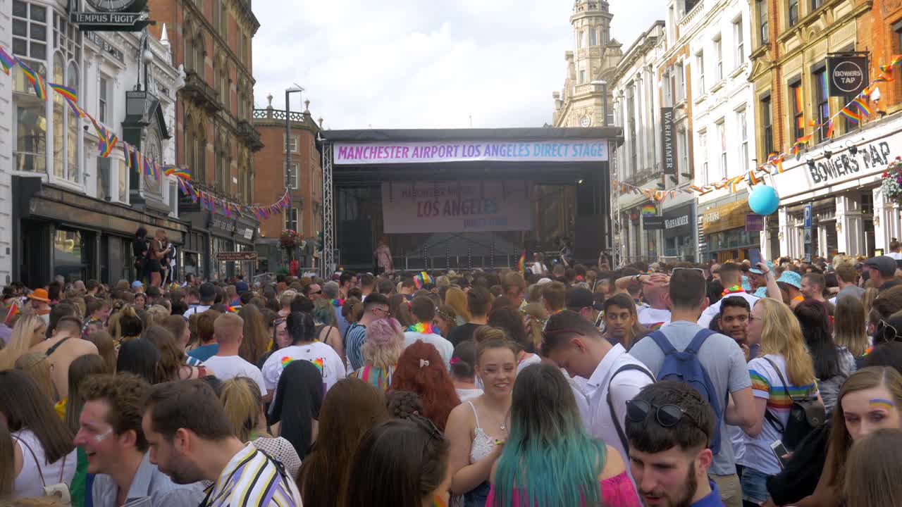 Leeds Pride LGBTQ Festival 2019 shot of the crowd in foreground dancing and waving flags with drag queens appearing on the stage dancing 4K 25p