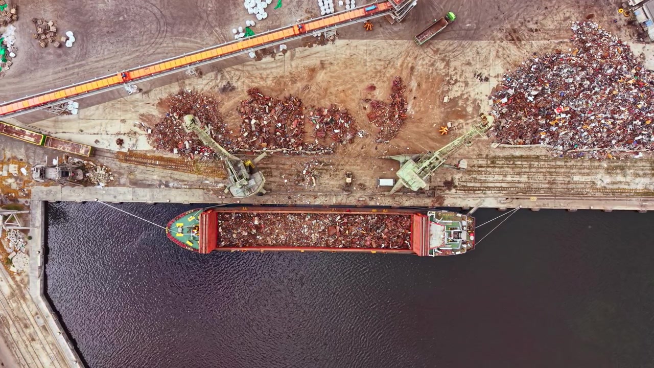 Gran buque de carga en un muelle cargando chatarra durante el día en una concurrida zona industrial.