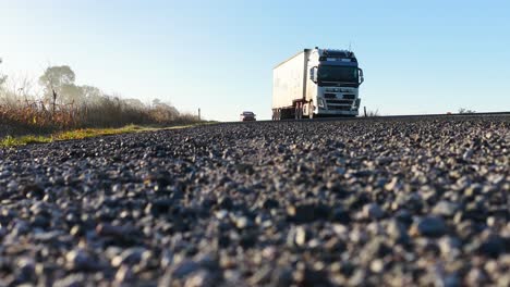 A large truck and red car travel along a rural gravel road in bright morning sunlight, captured from a low, ground-level perspective with a wide, static shot