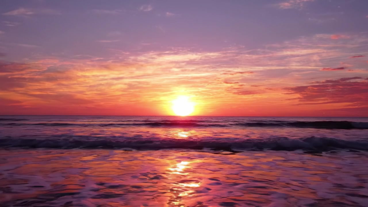En betagende solnedgang over det rolige hav, der oplyser himlen med levende nuancer af orange, pink og lilla, smukt reflekteret på de blidt rullende bølger