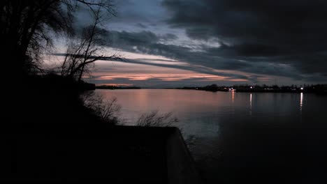 Pan-left-view-of-dark-clouds-over-still-river-waters-with-dramatic-clouds-and-colours-in-the-sunset-sky