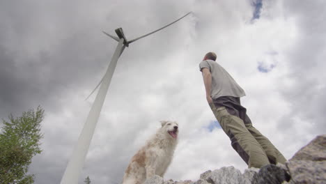 un hombre y su perro se paran debajo de una enorme turbina eólica