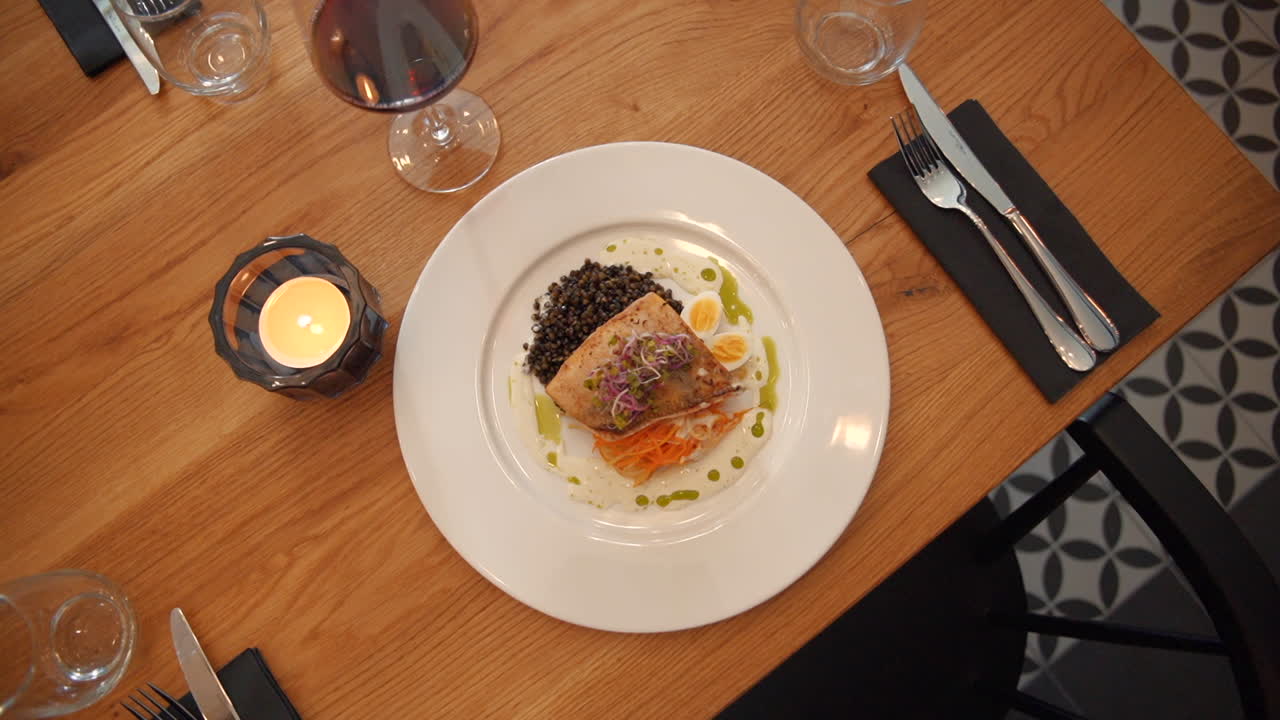 Beautifully presented salmon fillet with black lentils, pickled vegetables, garnished with a drizzle of herb oil served on white plate on wooden table. Fine dining perfection.