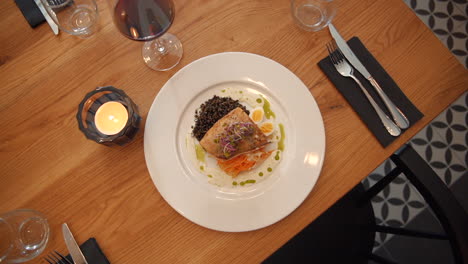 beautifully presented salmon fillet with black lentils, pickled vegetables, garnished with a drizzle of herb oil served on white plate on wooden table. fine dining perfection.