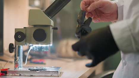 vista de primer plano de un microscopio con luces led que iluminan una placa de circuito en el banco de laboratorio, una persona con una bata de laboratorio y guantes negros se está preparando para examinar la placa