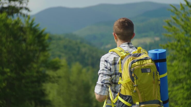 A Man With A Backpack Looks Ahead In Front Of Him Through Binoculars Back View Against The Backgroun