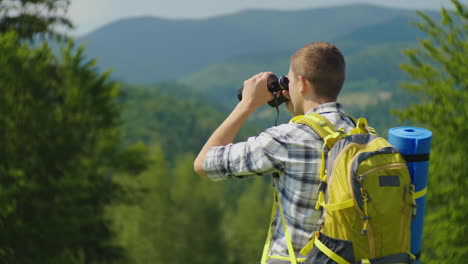 A-Man-With-A-Backpack-Looks-Ahead-In-Front-Of-Him-Through-Binoculars-Back-View-Against-The-Backgroun