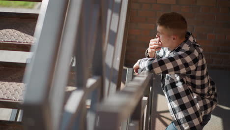 young student in checkered shirt and jeans leaning on iron railing smoking cigarette pack in hand cloth gently swaying in breeze sunlight filtering through metal stair casting shadow geometry