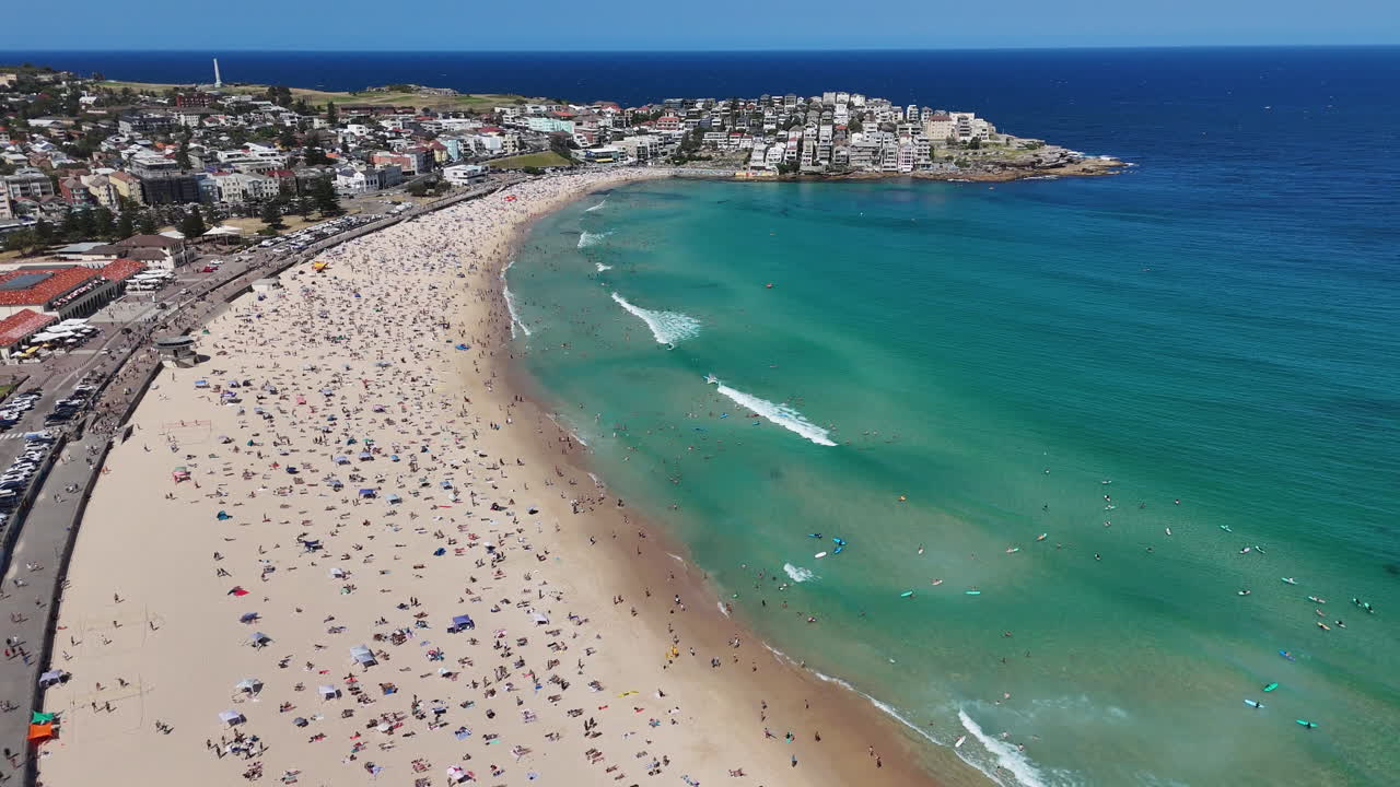 Wide drone shot of Bondi Beach with crowds soaking up the sun and swimming in the crystal-clear waters. Aerial shot, New South Wales.