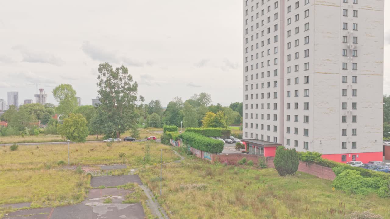Tall residential tower stands next to an overgrown vacant lot and surrounding greenery in Salford, England, captured in descending aerial shot