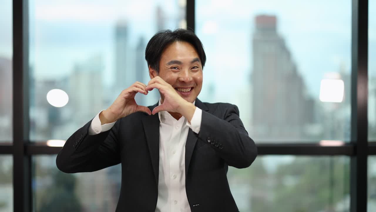 Confident businessman in suit forms heart shape with hands, smiling in bright modern office setting
