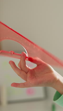 little girl pupil spins pink plastic square-ruler in fingers near cork-board in classroom closeup. schoolgirl plays with drawing tool resting in class