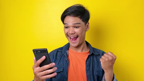 행복한 아시아의 젊은 남자가 휴대폰을 들고 승리하는 승리의 몸짓을 보여줍니다. 게임에서 승리합니다. 네, 남자가 주먹을  ⁇ 고 팔을 들어 올립니다. 노란색 바탕에 고립되어 있습니다.