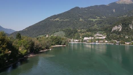 Gran-Lago-Con-Agua-Purificada-Que-Crea-Olas-Desde-La-Orilla-Del-Viento-En-La-Vista,-Casas-De-Una-Pequeña-Comunidad-De-Pueblo-En-Lo-Lejos,-Bajo-Enormes-Montañas-Cubiertas-De-árboles,-Cielo-Despejado,-Pájaro-Azul,-Escape-De-Drones