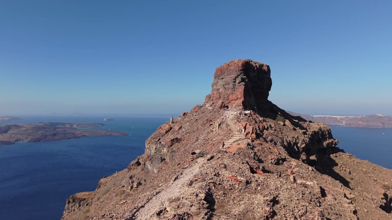 Orbita di drone attorno a Skaros Rock a Santorini con il vulcano sullo sfondo, catturando le spettacolari scogliere della caldera e l'iconico paesaggio insulare dell'Egeo.