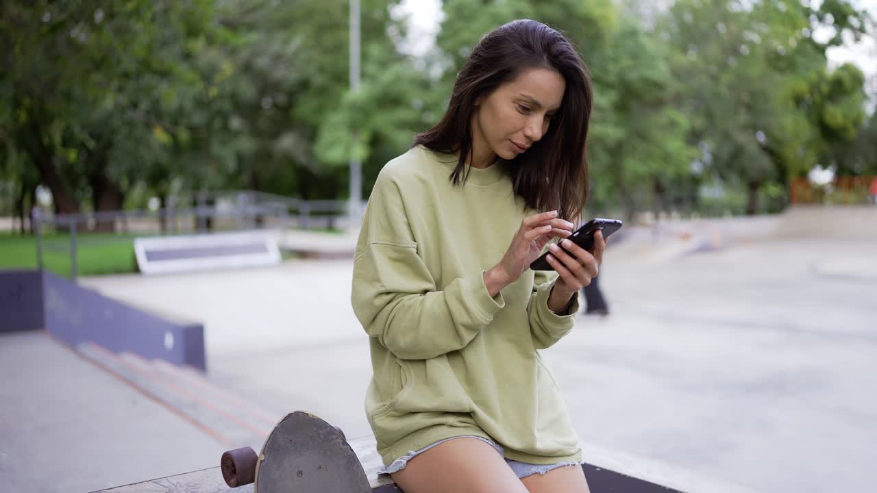 una chica morena con un suéter verde se sienta cerca de una patineta con el telón de fondo de un parque de patinaje y mensajes de texto en el teléfono. caminar