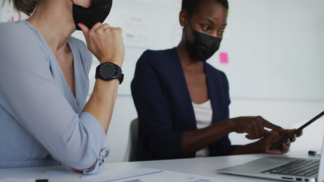 Two diverse female colleagues wearing face mask, sitting at desk and talking