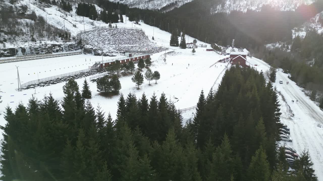 Dynamic 4K drone footage panning quickly over Vik Skisenter in Norway, showing cross-country skiers training on snowy tracks surrounded by pine forest and winter mountains