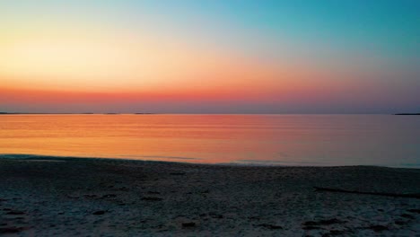 Precioso-Amanecer-En-El-Océano-Sobre-Una-Playa-De-Arena-Que-Proyecta-Coloridos-Reflejos-Rojos,-Naranjas,-Morados-Y-Amarillos-Sobre-Las-Tranquilas-Olas-Del-Mar.