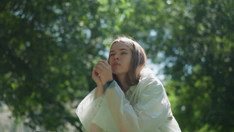 young woman in translucent raincoat thoughtfully removes her hood, resting on bicycle handlebars in dappled sunlight filtering through lush leaves