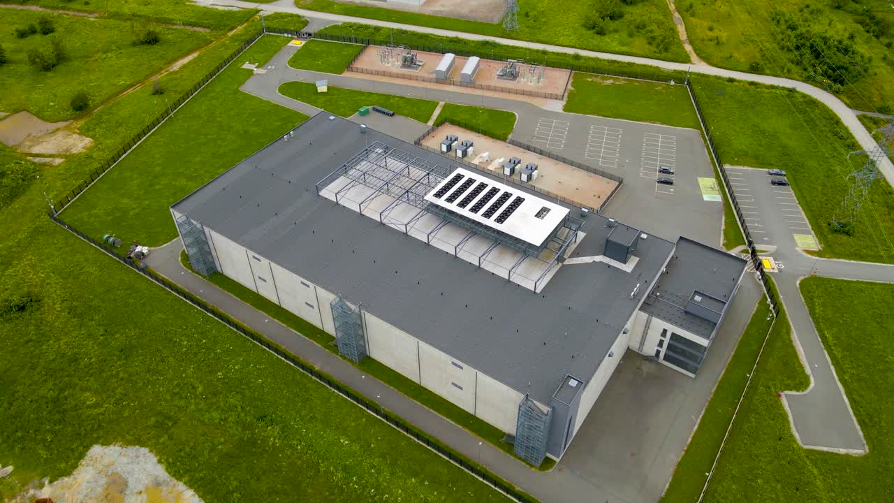 Aerial drone footage of a large and modern data centre or server housing complex warehouse during a cloudy day in europe countryside. A large electrical station in the background with electrical lines