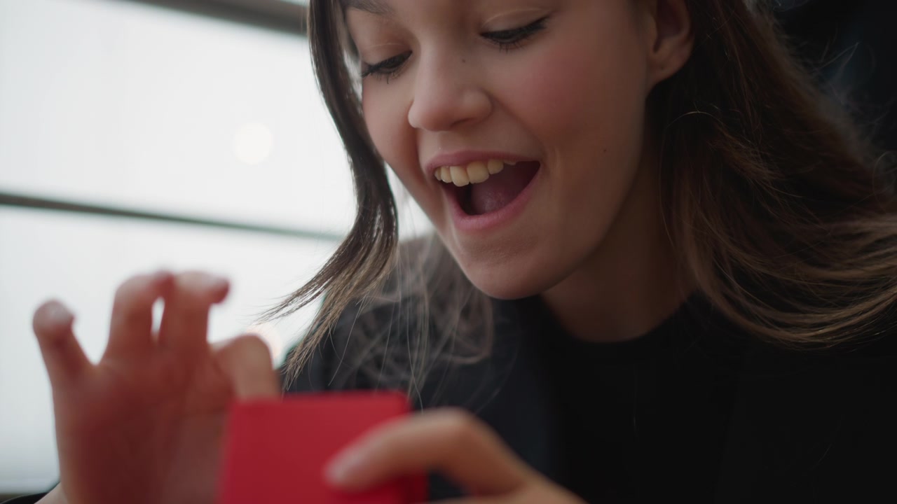 Young woman gently unties ribbon, lifts lid of red heart shaped gift box, and reveals romantic present inside while smiling with joyful surprise in warm soft light, with gift, heart, box, opening