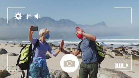 Smartphone-camera-interface-screen-over-caucasian-senior-couple-in-face-masks-on-coast-taking-selfie