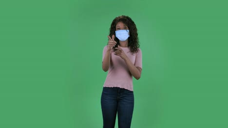 portrait of beautiful african american young woman in medical protective face mask looking at camera and pointing fingers to the side. burning brunette with wavy hair in jeans and a beige blouse on a green screen in the studio. health protection corona