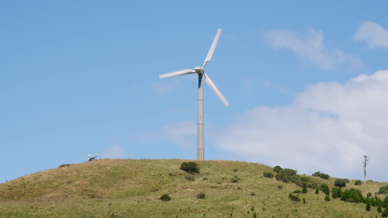 A single wind turbine generating power on top of a green hill with a clear sky in New Zealand