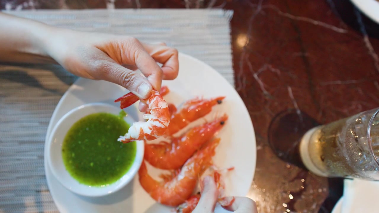 Vista dall'alto di mani che sgusciano gamberi su un piatto bianco con salsa verde piccante ai frutti di mare.