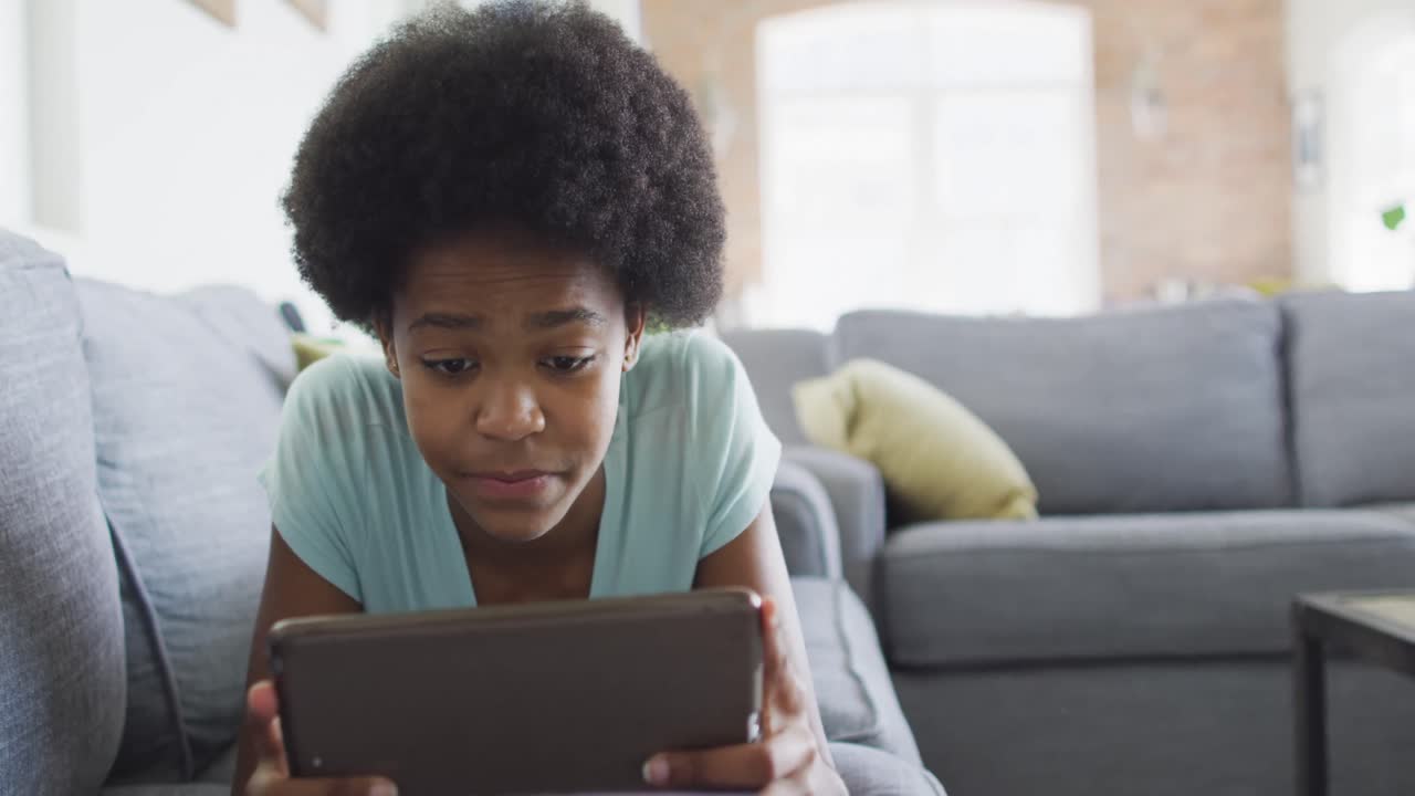Happy african american teenage girl lying on sofa, using tablet
