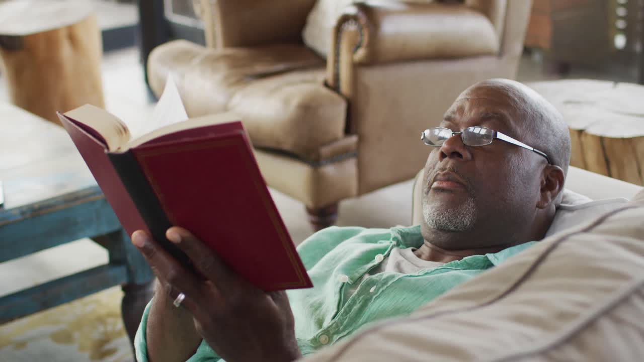 Glücklicher afroamerikanischer Senior entspannt sich, liegt auf dem Sofa und liest ein Buch.