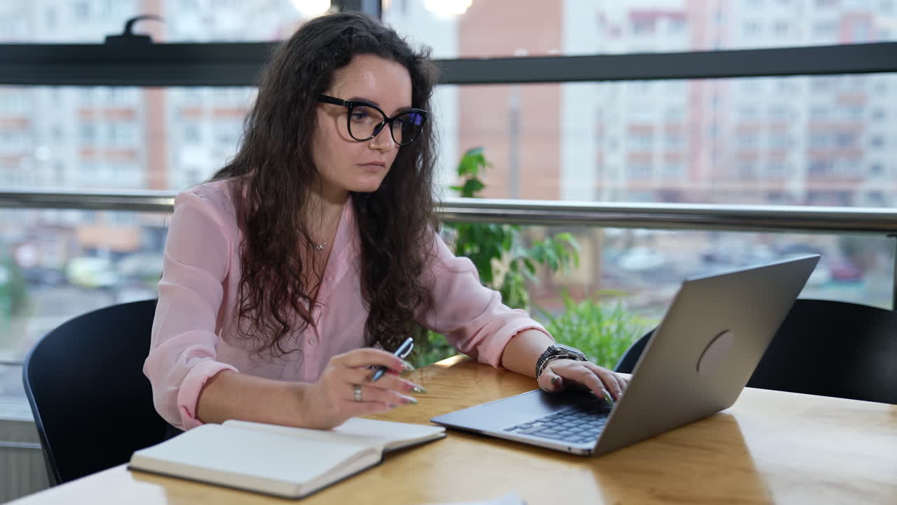 Businesswoman sitting at table in a spacious office, working with laptop and taking notes. Woman wearing glasses looking for information in computer.